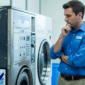 Man in blue shirt inspecting an old commercial washing machine, appearing deep in thought inside a laundry facility with Sudsy Vending Supplies branding.