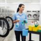 Laundry facility worker organizing cleaning tools and disinfectants beside industrial washers, showcasing essential janitorial supplies for commercial laundries.