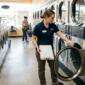 A laundromat attendant inspecting a commercial washing machine, holding a clipboard, clean and well-lit coin-operated laundry, rows of machines in the background